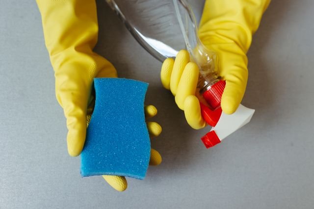 Hands wearing yellow rubber gloves holding blue cleaning sponge and spray bottle
